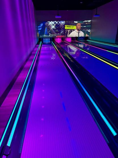 Modern bowling lanes illuminated with vibrant neon lighting in purple, blue, and pink hues. The lanes have LED strips along their edges that transition from blue to yellow to pink. Above the lanes, large TV screens are mounted on the wall showing sports content.