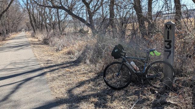 Blue and black Roubaix bicycle with Bar Mitts leaning against the concrete obelisk Chicago Great Western Railway milepost "M137" just off the asphalt Douglas State Trail looking Southeast. The trail is lined by bare trees on a sunny day.
