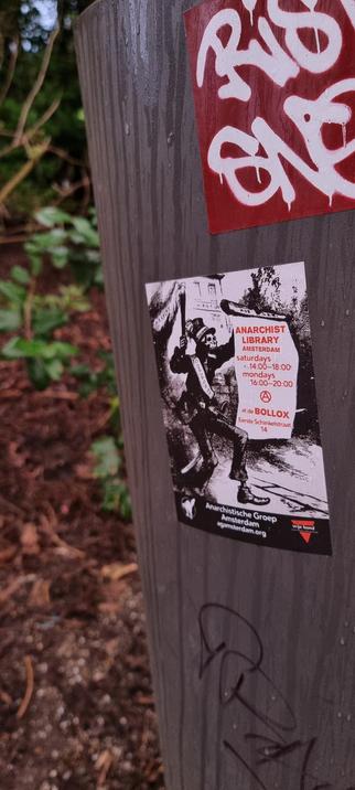A black and white sticker on the side of a trashcan in a park. It features a skelliton mayor with a torch burning with the word "anarchy" on it. They have a sash that says "communes" and they're holding a scroll that says:

MOB LAW. Anarchist Library Amsterdam saturdays 14:00-18:00 mondays 16:00-20:00 (anarchy symbol) at de BOLLOX Eerste Schinkelstraat 14

At the bottom of the sticker there is a black cat in halloween mode and it says "Anarchistische Groep Amsterdam agamsterdam.org" and in the bottom right corner is says "vrije bond anarchistische zelforganisatie"