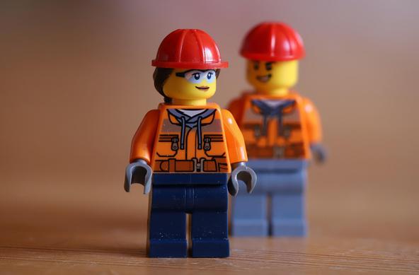 Man sieht zwei Lego Figuren, eine weibliche im Vordergrund und eine männliche im Hintergrund. Beide tragen einen roten Helm und ein oranges Oberteil, mit blauen Hosen. Die weibliche Figur ist scharf abgebildet, die männliche Figur verschwommen. Die Makro Aufnahme zeigt die beiden Figuren sehr gross und durch die Verwendung der Focus-Stacking Methode ist der Schärfebereich erweitert, so dass die weibliche Figur ganz scharf dargestellt ist.