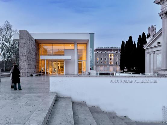 Vista esterna del Museo dell'Ara Pacis a Roma, progettato da Richard Meier. L'edificio moderno, con ampie vetrate e pareti bianche, contrasta con la struttura antica adiacente e la scalinata in travertino. In primo piano, una scritta incisa recita "ARA PACIS AUGUSTAE". Il cielo è crepuscolare.
