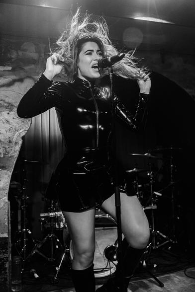 A black and white photo of a woman dressed in leather holding her hands up with her hair to the back as she performs.