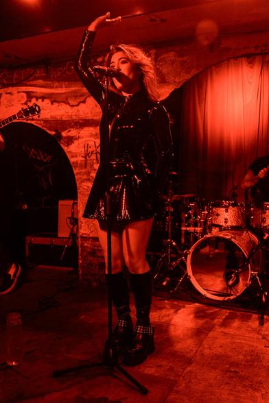 A black and white photo of a woman, full body shot, dressed in leather holding her hands up with her hair to the back as she performs. The room is awash with red light.