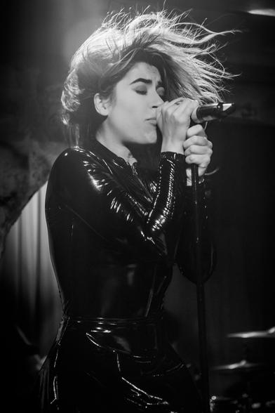 A black and white photo of a blonde woman dressed in leather singing into the microphone with her eyes closed.