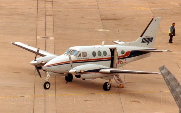 Side view of a grey, twin engined light aircraft parked facing to the left, with a door open in the rear fuselage hanging down to form steps.
There is a red and black stripe running along the body, repeated and inverted over the engines.
There are large black "CEGA"titles on the tail, and small "Cega Air Ambulance" titles under the cockpit windows.
The letters "WELL" are on the lower rear fuselage in red, part of the plane's registration.
Grey concrete apron fills the rest of the frame, with a man in a yellow hi-viz vest standing facing out of frame on the right in the background.