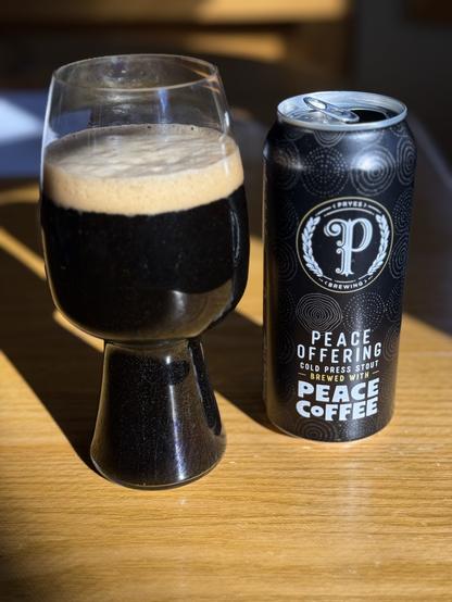 A brightly lit stout glass full of dark black beer sits on a wood table with a honey oak grain. The black can has white lettering with a large scripted “P” in a circle at the middle