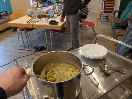 Foto vom Servierwagen mit einem Topf voller Käse-Lauch-Suppe, wo schon eine Kelle reingehalten wird. Im Hintergrund Tische mit Team-Mitgliedern.