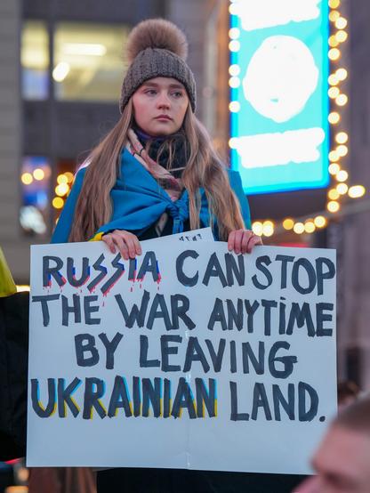 A young woman with long blonde hair, wearing a gray knit beanie with a pom-pom and a blue and yellow Ukrainian flag draped over her shoulders, stands in a public space at night holding a protest sign. The sign reads: 'RUSSIA CAN STOP THE WAR ANYTIME BY LEAVING UKRAINIAN LAND.' The word 'RUSSIA' is stylized with dripping red paint, resembling blood, and the words 'UKRAINIAN LAND' are highlighted in blue and yellow. Her expression is solemn and determined. Behind her, neon lights and blurred city buildings suggest an urban setting, possibly a well-known square or gathering place.