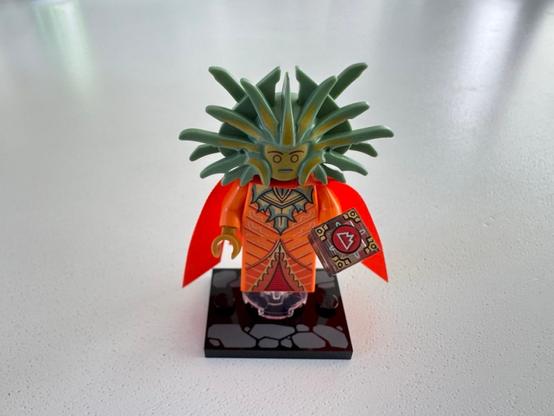 A Lego minifigure of the Lady of Pain from Planescape. Her body is mostly orange with dress lines. She has an orange fabric cape split into four segments. Her headdress of blades is yellow-green. She is holding transparent onesies with a red mountain glyph on them. The minifig is stacked on a transparent 2x2 cylinder on a 3x4 plate with cobblestone design.