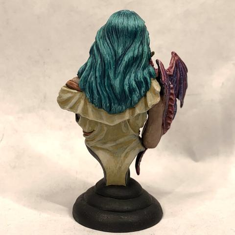 Bust of a woman holding a small pink and indigo dragon up to her face as if listening to it whisper. They look off to the right. She has long, silky teal hair, blue eyes, and wears a cream-colored frilly shirt. Over the shirt is a red leather harness with a gold medallion over her heart. She also wears a gold tiara.