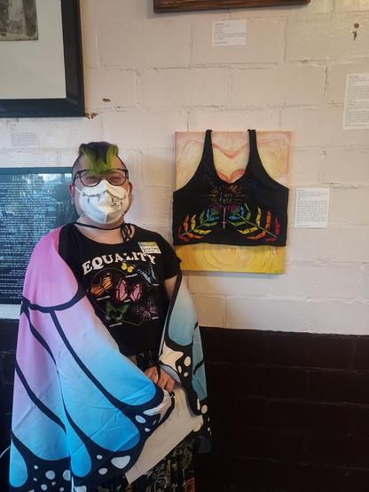 Me standing in a trans butterfly wings cloak in front of my binder embroidered and stretched over a canvas of my body with top surgery scars