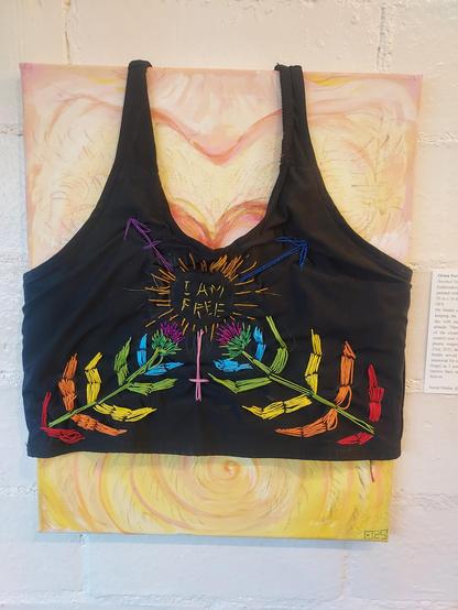 A binder embroidered to have rainbow thistle plant scars with "i am free" in a glowing trans symbol in the center of the chest. Its worn on a canvas that is my body with post op top surgery scars.