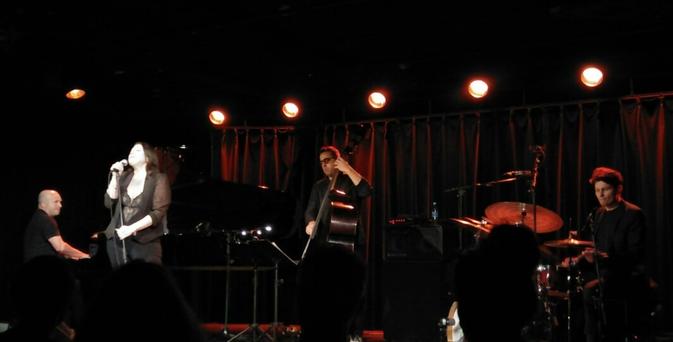 Foto von einer Bühne. Vier Musiker sind zu sehen, ein Pianist, eine Sängerin, ein Kontrabassist und ein Schlagzeuger. Das Licht ist abgedunkelt. Im Hintergrund ein dunkler Vorhang.