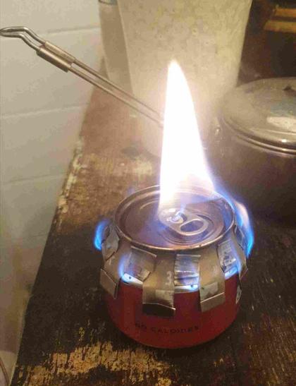 réchaud à alcool en canette en croisière, les flammes sortent par les trous sur le côté, le chapeau anti-condensation est posé dessus.
Les flammes s'échappent entre les pétales du chapeau et une flamme jaune semble sortir du chapeau, au milieu.
Les flammes sortant des trous sont bleues.