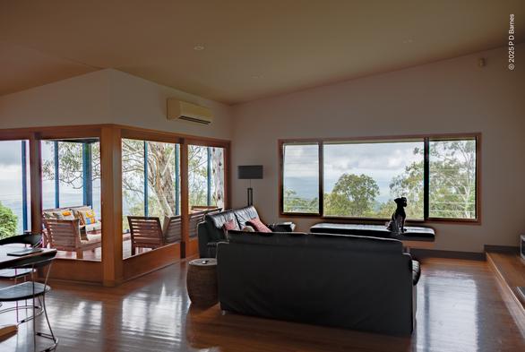 House interior with numerous sliding glass doors and picture windows looking out over a vista of trees and treetops. The interior has timber floors and frames, black leather furniture and white walls and ceiling. A dog is sitting on a low bench underneath the picture window, his silhouette against the trees outside.