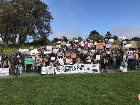 A couple of hundred people, some holding signs, stand on a low hill. The most prominent banner reads, "Protect Our Parks & Rangers."