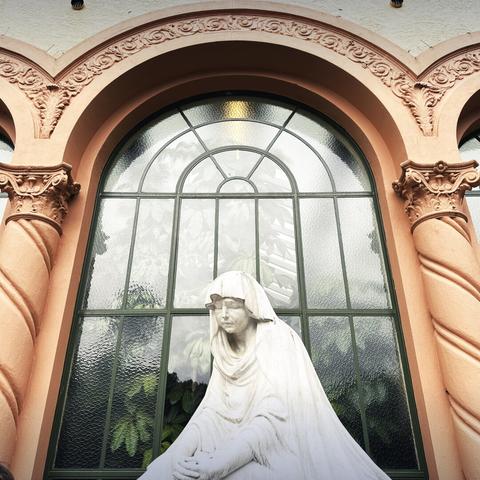 A statue of a sad-looking shrouded figure, in front of a  curved window that shows the reflection of some clouds.