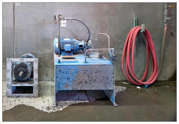 An industrial setup with machinery and equipment. On the left side, there is a large blue motor mounted on a metal table, which shows signs of wear and paint chipping. Attached to it is possibly a pump or compressor of some sort, connected via piping that might be part of a fluid handling system. Next to the motor setup, a detached fan component is visible, also appearing worn and used.

On the right, there is a red hose coiled and hanging from a hook on the concrete wall. This hose might be used for air or water delivery as part of the industrial processes in this setting. The wall and floor also show signs of use, with stains and residues, likely from the industrial operations in this area. There is also some minor debris on the floor, including what looks like small metal or concrete pieces. 

Overall, the setting suggests a mechanical or maintenance workshop environment.