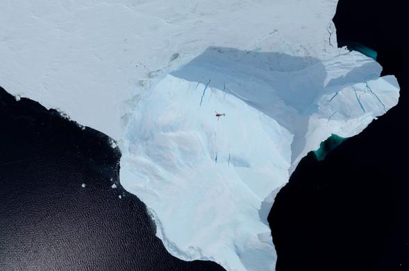 A photograph of a helicopter flying over an iceberg, shot with a downward looking camera in another helicopter flying above