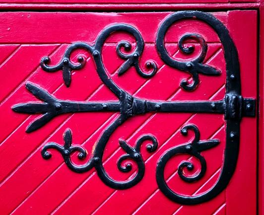 A large, ornate iron hinge, painted black, on a bright red church door.