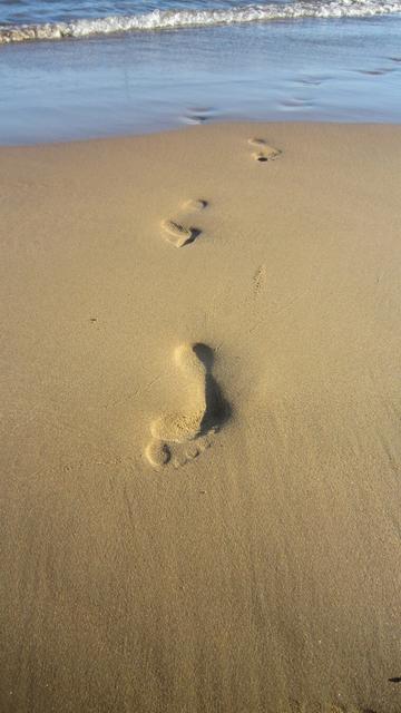 A close-up, slightly angled shot captures a section of a sandy beach. The focus is on a series of human footprints imprinted in the light brown, textured sand. The footprints are arranged in a diagonal line, moving from the bottom right corner of the image towards the upper left, indicating a person walking away from the viewer and away from the ocean. The closest footprint, located at the bottom right, is large and clear, showing the distinct shape of a human foot with toes and heel. The sand around the footprints is slightly disturbed, showing the texture and the imprint's depth. As the footprints recede into the distance, they become smaller and less defined. In the upper left portion of the image, a thin strip of blue ocean water is visible, with a gentle wave breaking along the shoreline. The transition between the wet sand near the water and the dry sand where the footprints are is subtle.
