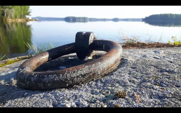 Kallio, johon kiinnitetty paksu rautarengas puutavaranippujen kiinnittämistä varten. Taustalla tyyni järvi ja saaria