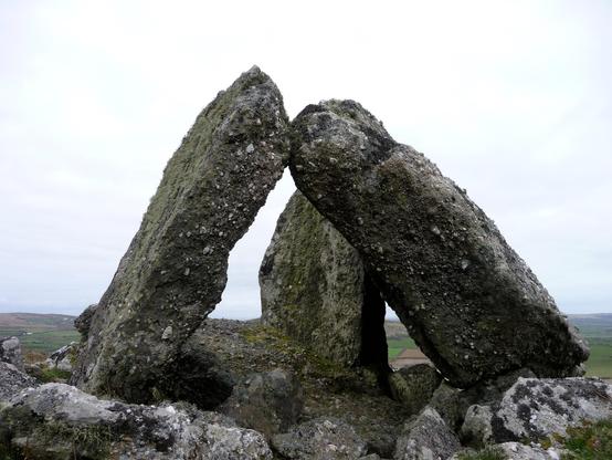 Three large upright stones of a pebbly conglomerate stone. The two stones on the left are the remaining supporting orthostats of a chamber. The stone on the right is a large slab which would have formed the capstone. The chamber is surrounded by smaller stone blocks, which remain from a surrounding or covering cairn. It's a grey, overcast day.
