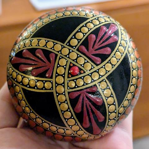 A round, dome-shaped plaster rock painted black then decorated with a three-pointed celtic knot called a "triquetra". The knot's lines are drawn with gold pen and filled in with metallic gold dots. The interior of each of the three points has a dark red swoosh design inside. There's a bright metallic red red dot at the centre.