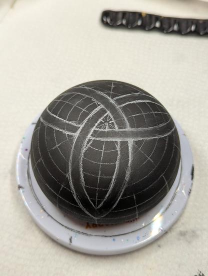 A photo of the stone before it's decorated, simply painted black. It's sitting on a small turntable, and is covered in chalky white grid lines, with a sketchy triquetra drawn using the grid lines as a guide. Other than the grid lines, the triquetra was free-handed.