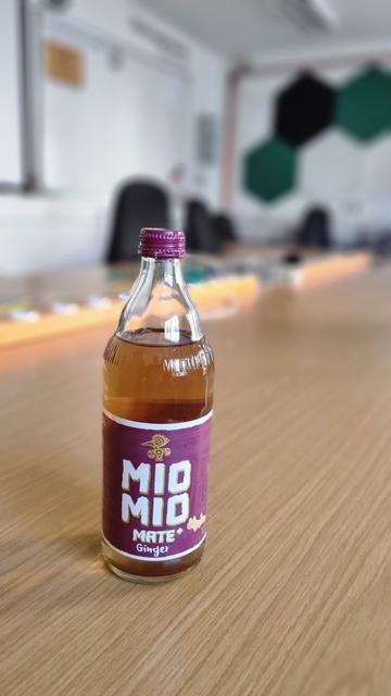 Foto einer Flasche Mio Mio Mate+ Ginger. Im Hintergrund sind unscharf Steckdosenleisten, Bürostühle und farbige Muster an einer Wand zu erkennen.