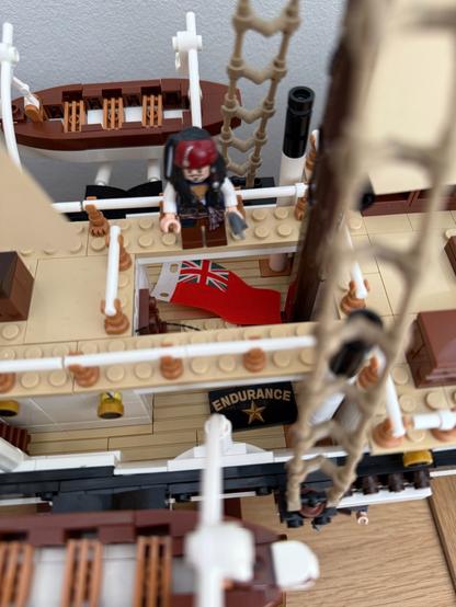 A detailed model of a ship featuring a pirate minifigure on deck. The ship's interior includes a flag with the Union Jack and a banner that reads "ENDURANCE." There are also rowboats and rigging visible in the scene.