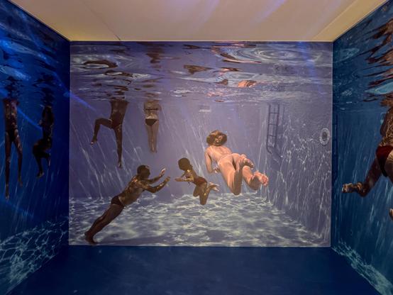 Una stanza progettata per assomigliare a una scena subacquea. Le pareti e potenzialmente il pavimento sono ricoperte da grandi immagini di persone che nuotano sott'acqua, creando un'esperienza coinvolgente. I riflessi e l'illuminazione imitano come apparirebbero le cose sott'acqua, migliorando l'effetto realistico.