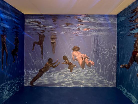 Una stanza progettata per assomigliare a una scena subacquea. Le pareti e potenzialmente il pavimento sono ricoperte da grandi immagini di persone che nuotano sott'acqua, creando un'esperienza coinvolgente. I riflessi e l'illuminazione imitano come apparirebbero le cose sott'acqua, migliorando l'effetto realistico.