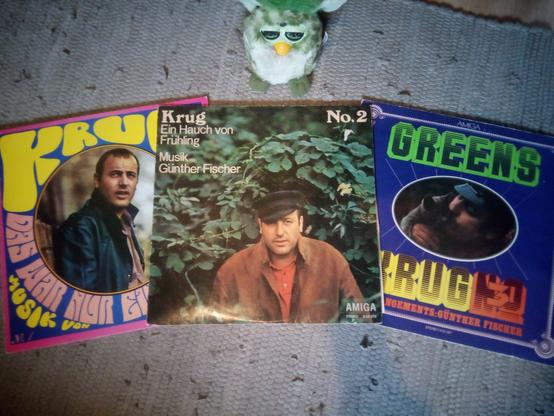 Auf einem grauen Teppich auf dem Boden liegen drei Schallplatten von Manfred Krug (und Günther Fischer). Von links nach rechts: "Das war nur ein Moment", "Ein Hauch von Frühling" und "Greens". Darüber steht ein grüner Furby und blickt auf die Platten.