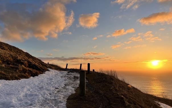 A winter coastal sunrise from Signal Hill National Historic Site. An icy snow covered path follows an ocean cliff edge in the left of the image, marked by short wooden posts.  The right side of the image shows  a yellow orange sun reflecting on the open the ocean toward the horizon, giving an orange glow to snow patches on the cliff side. The blue sky has puffy clouds tinged orange and purple, becoming more dense near the horizon. [St. John’s (NL, Canada) March 2025]