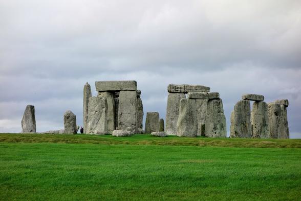Stonehenge stands against a cloudy sky, its ancient stones silhouetted on the horizon. The iconic monument is composed of large, rough-hewn stones, some upright and others lying on the green field. The stones are weathered and grey, showing signs of age. The green grass in the foreground is lush and vibrant, contrasting with the grey stones and the overcast sky. The sky is filled with grey clouds, suggesting a cool or overcast day. The overall scene is serene and evocative, capturing the timeless mystery of Stonehenge.