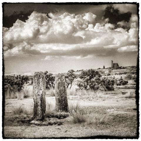 A toned black and white infrared photo of two standing stones together on moorland, with a ruined building on the skyline.
