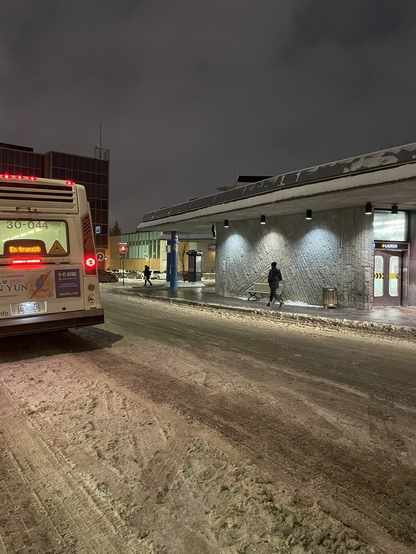 A street at night. There is an entrance to a subway station and a bus parked across the street. Someone is walking on the sidewalk.