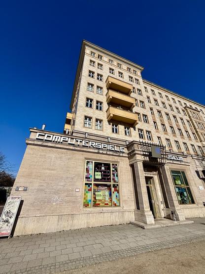 The image shows the exterior of the Computerspielemuseum in Berlin, a museum dedicated to the history of video games. The building has a classic stone facade with large windows decorated with pixel art and gaming-related imagery. The entrance is framed by a retro-style sign displaying the museum’s name in bold capital letters. The sky above is a clear and vibrant blue, adding to the inviting atmosphere. Graffiti is visible on the lower part of the wall, giving the scene an urban touch.