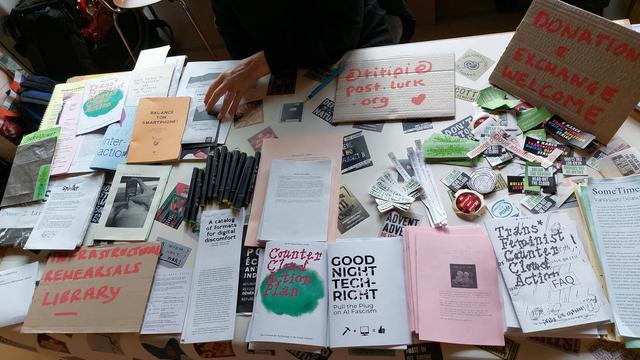 A table filled with booklets, zines, pens, stickers. A sign reads: Infrastructural rehearsals library.