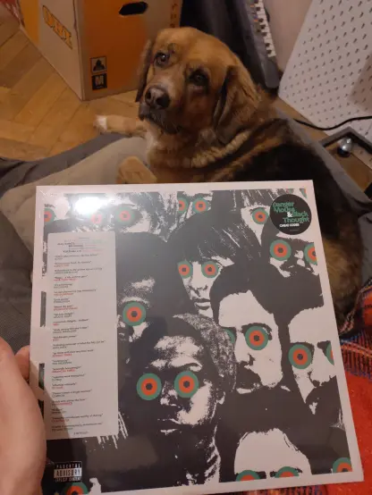 A vinyl album held in hand, still wrapped in plastic. The album cover features a bunch of black and white stylized heads, some of famous people. All have black, red and green circles instead of eyes.
A brownish dog is lying in its bed in the background and looking at the camera.
There is a moving box and some other stuff behind the dog.