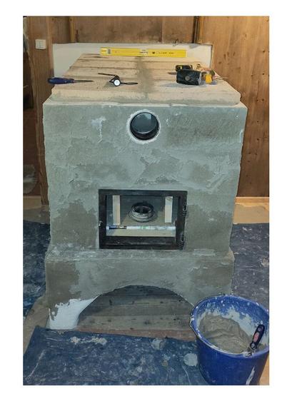 Oven ceiling closed with fireclay plate bricks. Old oven door frame refitted in front of the ofen entrance. Blue bucket with fireclay mortar and spatula inside stands on a blue plastic bag that covers the floor.
Tools such as a spirit level, a spatula, a flashlight, a tape measure, a rubber hammer and brass brush lying on the top of the oven.
 A old wooden paneling is visible on the left, top and right edges of the photo.
