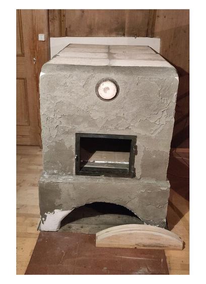 Oven ceiling closed with fireclay plate bricks. Old oven door frame refitted in front of the ofen entrance. On old ceramic lid of the claning opening is fitted above the oven door. Terracotta tiles visible in front of the ofen with two wooden templates for the round arch on it. A old wooden paneling is visible on the left, top and right edges of the photo. A wooden parquet floor is visible to the left and right of the stove and a wooden sliding door with a light switch in its frame on the left side of the oven.