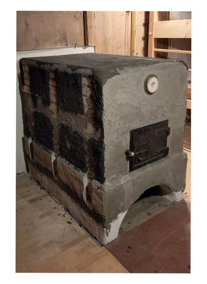 Gray plastered oven sideview with oven sides prepared for adding a support frame.
Old oven door fitted in front of the ofen entrance. The old lid of the claning opening fitted above the oven door. Terracotta tiles visible in front of the ofen with two wooden templates for the round arch on it. A old wooden paneling is visible on the left, top and right edges of the photo. A wooden parquet floor is visible to the left and right of the stove and a wooden sliding door with a light switch in its frame on the left side of the oven.