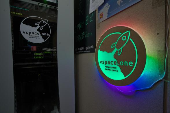 Ein Foto von unserem Serverrack und dem beleuchteten vspace.one Lasercut Logo Schild im Gang. Auf der Glastür vom Rack, durch die man eine handvoll Server und Switches erkennen kann, ist ein weißes vspace.one Logo drauf. Das braune runde Schild rechts davon ist am Rand in Regenbogenfarben beleuchtet und das Logo auf der Fläche in Grün. Im Hintergrund sind noch einige Poster unter anderem von der GPN22 zu erkennen.