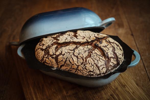 Farbiges Querformatbild: Ein Sauerteigbrot in einer blauen Form. Der Deckel, ebenfalls blau ist hintendran gelegt. Das Brot zeigt tiefe Risse und ist mit Mehl bestrichen.