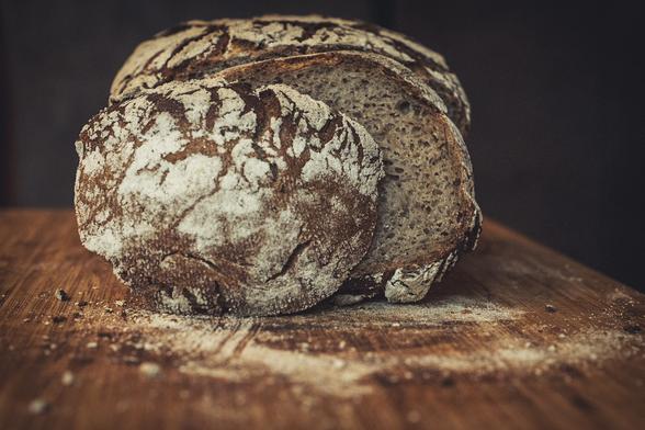 Querformat, Farbe. Das Brot ist aus einer tieferen Position heraus fotografiert. Es sind zwei Scheiben abgeschnitten. Die Vorderste  ist der Anschnitt, der stark mit Mehl bestrichen ist. Man sieht ausserdem das Mehl auf dem Holzbrett, das einen starken Schärfeverlauf zeigt. Der hintere Bereich ist dunkel.