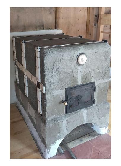 Gray plastered oven sideview with oven sides with iron support frame.
Old oven door fitted in front of thae ofen entrance. The old lid of the claning opening fitted above the oven door. Terracotta tiles visible in front of the ofen with two wooden templates for the round arch on it. A old wooden paneling is visible on the left, top and right edges of the photo. A wooden parquet floor is visible to the left and right of the stove and a wooden sliding door with a light switch in its frame on the left side of the oven.