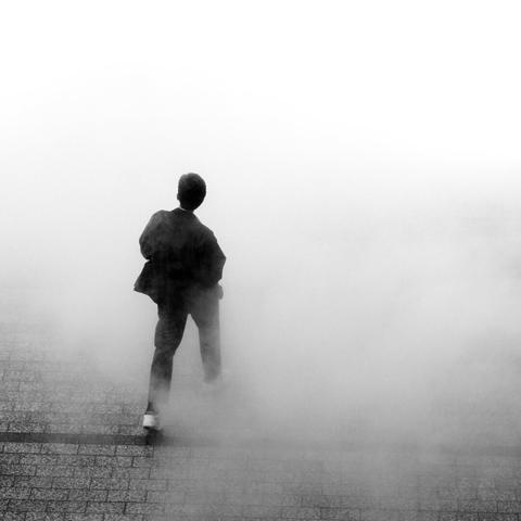 The image depicts a person standing or walking on a pavement covered in mist or fog. The scene is monochromatic, with varying shades of gray and white, creating a mysterious or ethereal atmosphere.