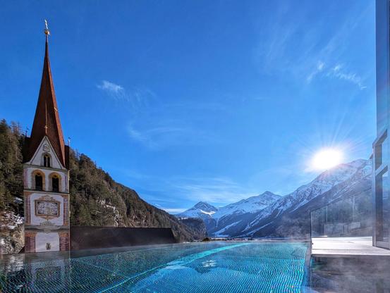 Infinity-Pool mit Aussicht auf die Kirche mit teich verziertem Turm und die verschneiten Berge rundum. Die Sonne verschwindet bald hinter den Bergen.